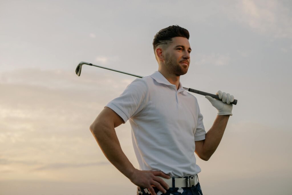 A confident male golfer in a white polo shirt posing with a golf club at sunset.