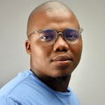 A confident young man wearing glasses and a blue shirt against a neutral background.
