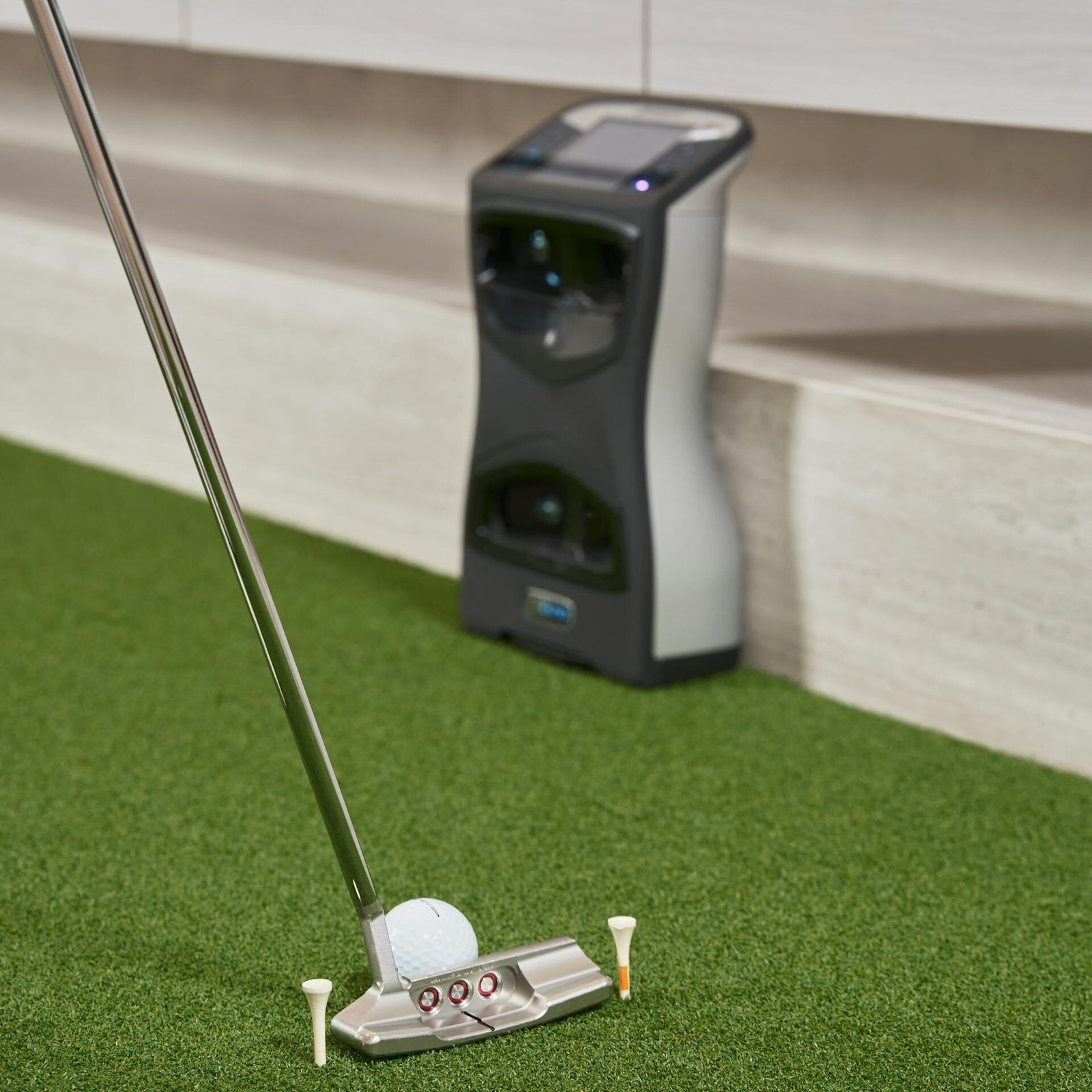 Indoor setup for practicing golf putting with a high-tech sensor and putter on artificial turf.